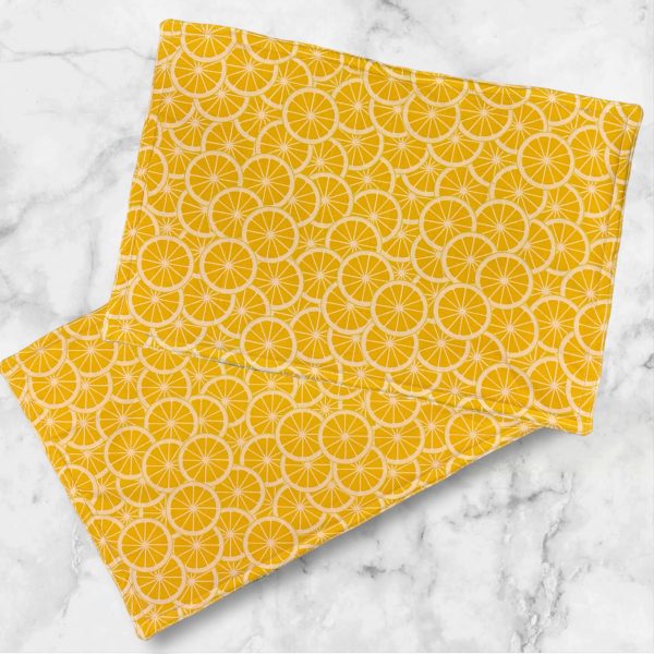 A set of two 19x13 inch fabric placemats, featuring a diagonal patchwork design with green and yellow patterns on one side and a bright lemon slice print on the other, laid on a marble background.