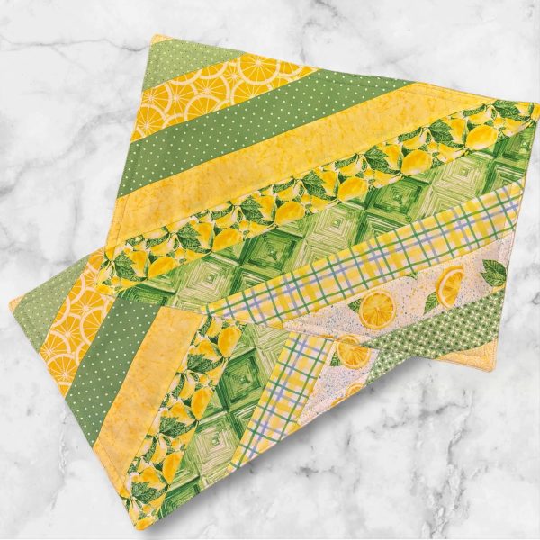 A set of two 19x13 inch fabric placemats, featuring a diagonal patchwork design with green and yellow patterns on one side and a bright lemon slice print on the other, laid on a marble background.