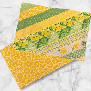 A set of two 19x13 inch fabric placemats, featuring a diagonal patchwork design with green and yellow patterns on one side and a bright lemon slice print on the other, laid on a marble background.