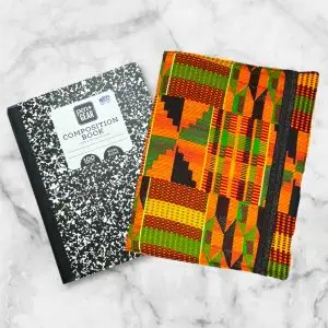 Kente-inspired composition notebook cover featuring bold oranges, greens, blacks, and yellows in intricate geometric patterns with an elastic band.