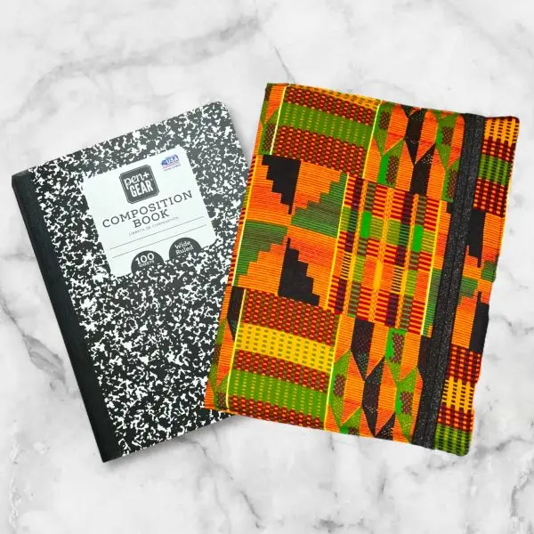 Kente-inspired composition notebook cover featuring bold oranges, greens, blacks, and yellows in intricate geometric patterns with an elastic band.