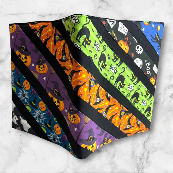 A composition notebook cover with diagonal stripes featuring Halloween-themed fabrics, including black cats, pumpkins, witches' hats, and ghosts in vibrant colors like orange, green, purple, and blue. The cover is displayed next to a standard composition notebook.