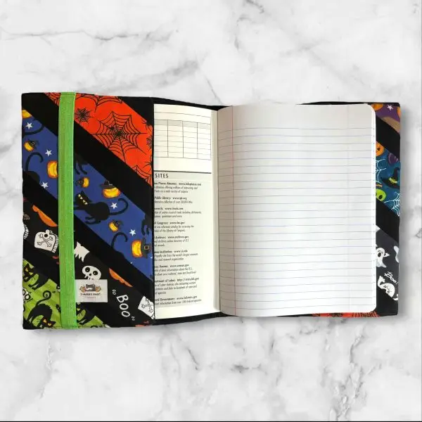 A composition notebook cover with diagonal stripes featuring Halloween-themed fabrics, including black cats, pumpkins, witches' hats, and ghosts in vibrant colors like orange, green, purple, and blue. The cover is displayed next to a standard composition notebook.