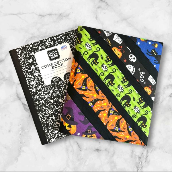 A composition notebook cover with diagonal stripes featuring Halloween-themed fabrics, including black cats, pumpkins, witches' hats, and ghosts in vibrant colors like orange, green, purple, and blue. The cover is displayed next to a standard composition notebook.