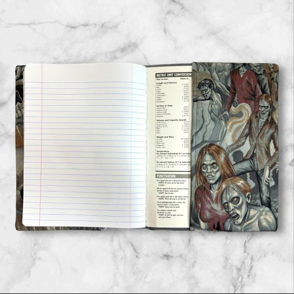 A composition notebook cover showcasing a detailed illustration of a crowd of zombies in various eerie poses. The cover has a gray elastic band securing it closed and is displayed next to a standard composition notebook.