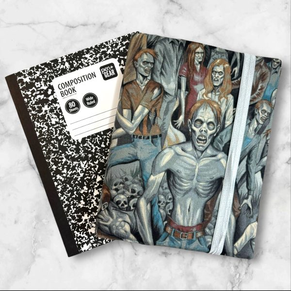 A composition notebook cover showcasing a detailed illustration of a crowd of zombies in various eerie poses. The cover has a gray elastic band securing it closed and is displayed next to a standard composition notebook.