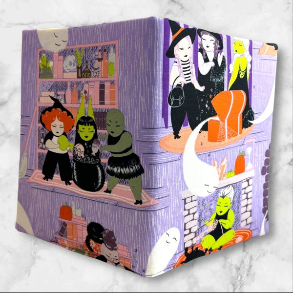 A composition notebook cover with a fabric design showcasing witches, ghosts, and Halloween-themed scenes in bright colors, predominantly purple, green, and orange. The cover is displayed with a standard composition notebook.