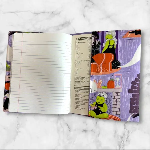 A composition notebook cover with a fabric design showcasing witches, ghosts, and Halloween-themed scenes in bright colors, predominantly purple, green, and orange. The cover is displayed with a standard composition notebook.