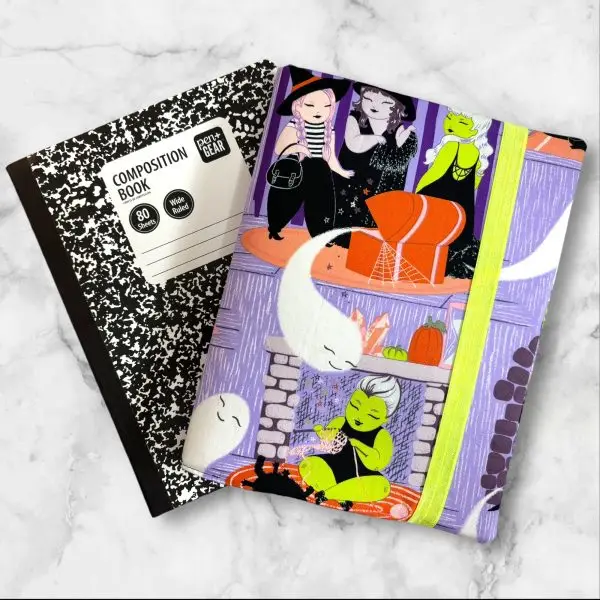 A composition notebook cover with a fabric design showcasing witches, ghosts, and Halloween-themed scenes in bright colors, predominantly purple, green, and orange. The cover is displayed with a standard composition notebook.