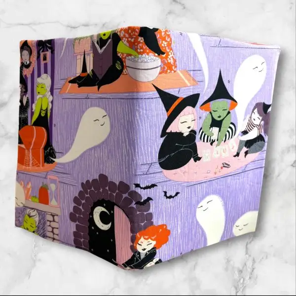 A composition notebook cover featuring a whimsical witch-themed fabric with illustrations of witches, ghosts, and playful scenes in vibrant colors against a lavender background. The cover has a neon green elastic band and is shown next to a standard composition notebook.