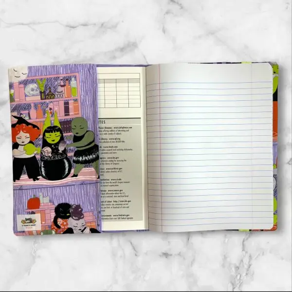 A composition notebook cover featuring a whimsical witch-themed fabric with illustrations of witches, ghosts, and playful scenes in vibrant colors against a lavender background. The cover has a neon green elastic band and is shown next to a standard composition notebook.