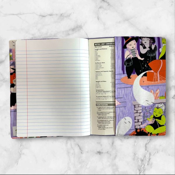 A composition notebook cover featuring a whimsical witch-themed fabric with illustrations of witches, ghosts, and playful scenes in vibrant colors against a lavender background. The cover has a neon green elastic band and is shown next to a standard composition notebook.