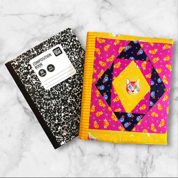 Quilted composition notebook cover with a pink and yellow design, showcasing a playful Cheshire Cat print surrounded by bright floral patterns.