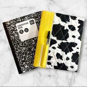 Quilted composition notebook cover featuring a black and white cow print with a bright yellow accent, complete with a pen holder on the side.