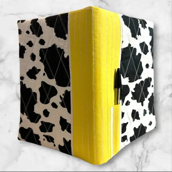 Quilted composition notebook cover featuring a black and white cow print with a bright yellow accent, complete with a pen holder on the side.