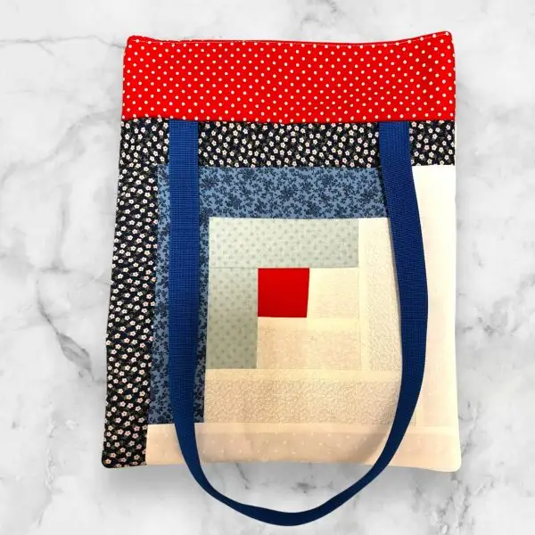 A handmade quilted tote bag featuring a blue and red log cabin front design with floral accents and a blue patchwork-patterned back.