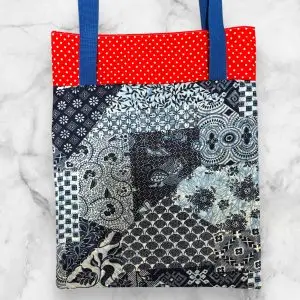 A handmade quilted tote bag featuring a blue and red log cabin front design with floral accents and a blue patchwork-patterned back.