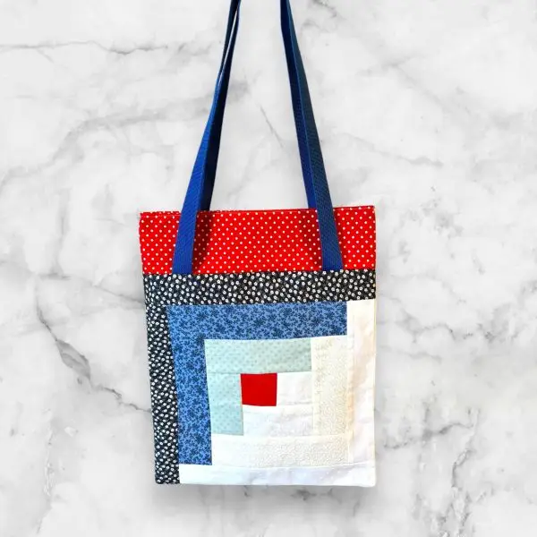 A handmade quilted tote bag featuring a blue and red log cabin front design with floral accents and a blue patchwork-patterned back.