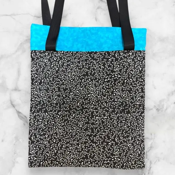 A handmade quilt block tote bag featuring a patchwork front with black-and-white patterns, a turquoise accent, and a vine-print back.